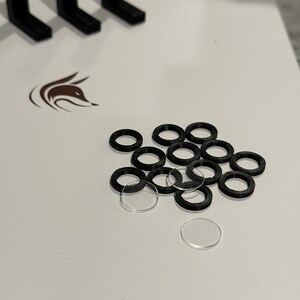 以下が含まれることがあります： 白い表面に、黒と透明なプラスチック製のリングが積み重ねられている様子のクローズアップ。リングはさまざまなサイズで、ランダムなパターンで配置されています。