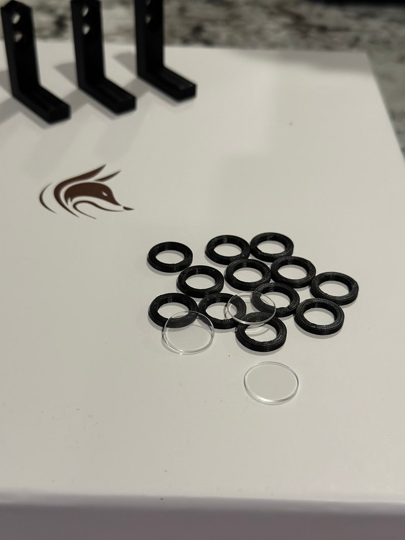以下が含まれることがあります： 白い表面に、黒と透明なプラスチック製のリングが積み重ねられている様子のクローズアップ。リングはさまざまなサイズで、ランダムなパターンで配置されています。