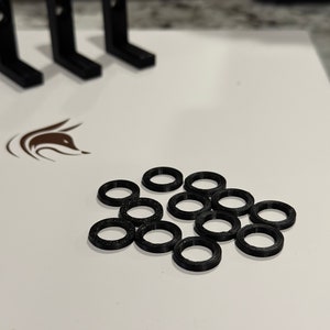 以下が含まれることがあります： 白い表面に、3Dプリンター用の黒いプラスチックリングが18個散らばっている。