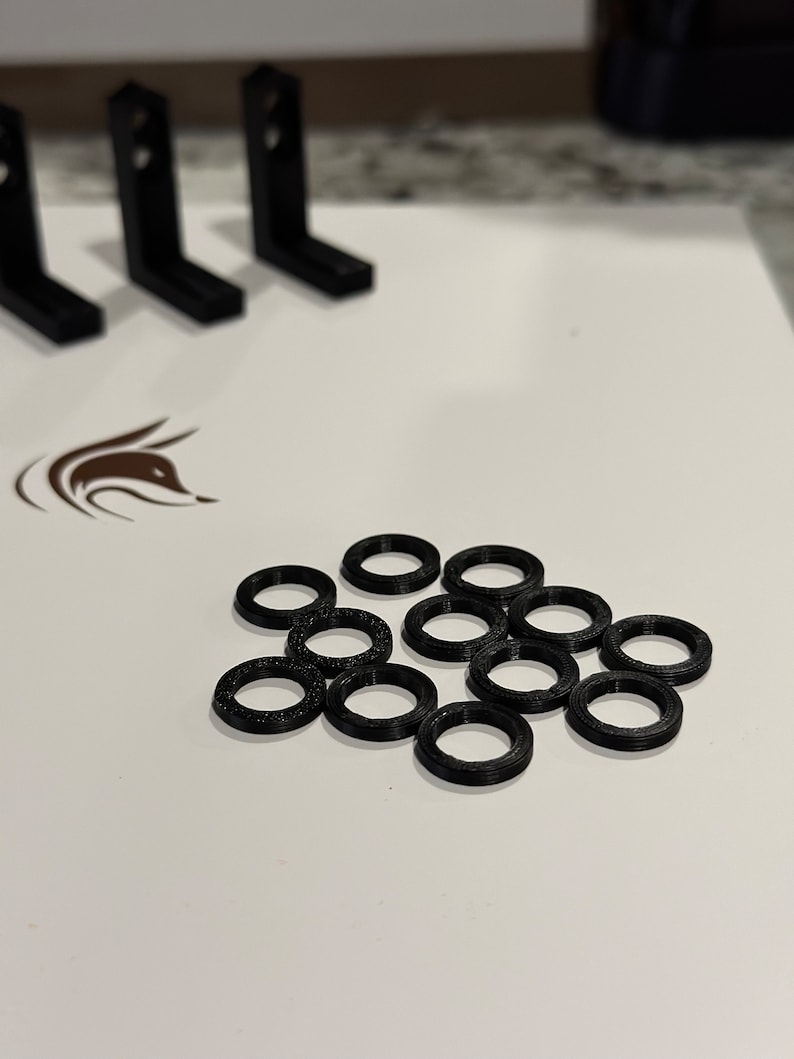 以下が含まれることがあります： 白い表面に、3Dプリンター用の黒いプラスチックリングが18個散らばっている。