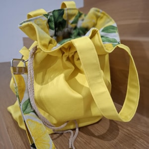 Puede incluir: Un bolso amarillo brillante con cordón y una correa con temática de limón. El bolso tiene un exterior amarillo y un estampado de limón y hojas verdes en el forro interior. Tiene dos asas amarillas y un cierre de cordón.