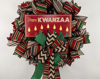 May include: A red, green, and black Kwanzaa wreath with a sign that reads "Happy Kwanzaa" and a depiction of seven candles. The wreath is decorated with a bow made of green, black, and gold ribbon.