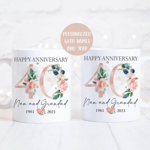 May include: Two white mugs with a floral design and the number 40 in rose gold. The text "Happy Anniversary" is at the top of each mug. The text "Nan and Grandad" is below the number 40. The years 1983 and 2023 are below the names.
