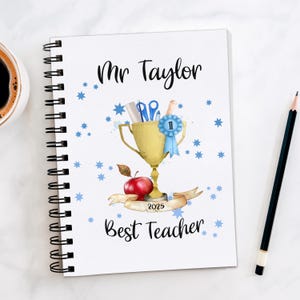 May include: A spiral-bound notebook with the text "Mr Taylor Best Teacher 2025." The cover design includes a gold trophy, blue stars, a red apple, and school supplies. A pencil and a cup of coffee are on the side.