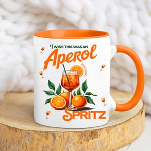 May include: White ceramic mug with an orange interior and handle. The mug features the text "I wish this was an Aperol Spritz" with a graphic of the cocktail, oranges, and leaves. The name "Hannah" is also on the mug.