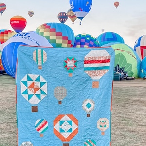 May include: A blue quilt with a patchwork design featuring hot air balloons in various colours and patterns. The quilt is displayed in front of a field with hot air balloons in the background.
