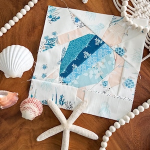May include: A square fabric block with a patchwork design in shades of blue, white, and pink, featuring floral and sea turtle patterns. A starfish, seashells, and wooden beads surround the block, creating a coastal theme.