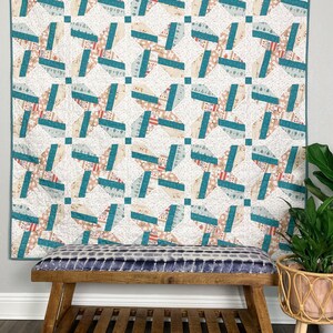 Puede incluir: Una colcha de color turquesa y crema con un patrón geométrico cuelga de una pared sobre un banco de madera con un cojín de estampado azul y blanco.