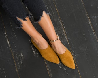 Pode incluir: Um par de sapatos de camurça marrom com bico fino. Os sapatos são usados por uma pessoa que está vestindo jeans preto e uma pulseira de conchas.