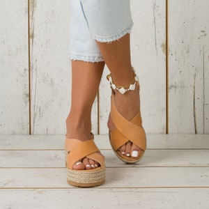 May include: A pair of tan leather sandals with a criss-cross strap design and a platform sole. The sandals have a white ankle bracelet with white beads.