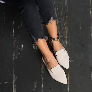 Könnte beinhalten: Ein Paar hellbeige Slipper aus Wildleder mit spitz zulaufenden Zehen. Die Schuhe werden von einer Person getragen, die eine dunkle Jeans trägt.