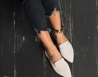 Pode incluir: Um par de sapatos de camurça bege claro com bico fino. Os sapatos são usados por uma pessoa que usa jeans escuros.