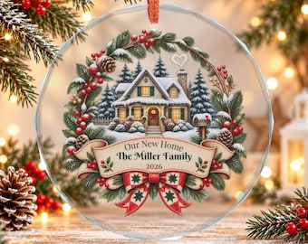 May include: A circular glass ornament featuring a winter scene with a snow-covered house, pine trees, and a wreath. A banner across the bottom reads Our New Home, The Miller Family, 2026, accented by a red and green patterned bow.