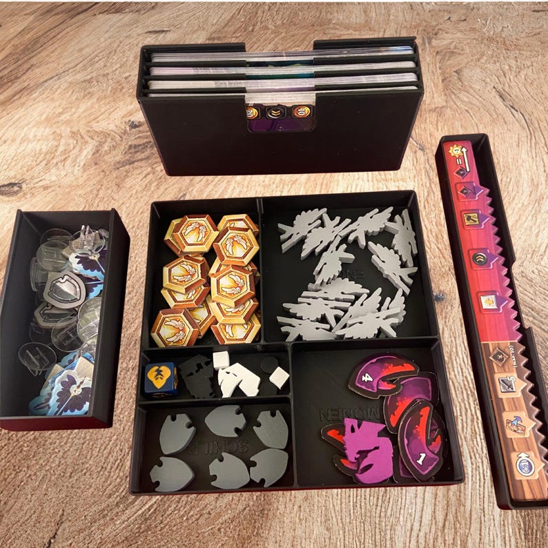 May include: A black plastic board game organizer set featuring a card holder, a long tray with a vertical red and orange track, and a multi-compartment tray containing hexagonal gold tokens, grey wing-shaped pieces, small white cubes, and purple tokens.