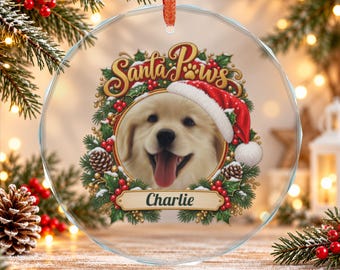 May include: A round, clear glass Christmas ornament featuring a golden retriever puppy wearing a red Santa hat. The puppy is framed by a wreath of holly, pinecones, and red berries. The text reads Santa Paws and the name Charlie.