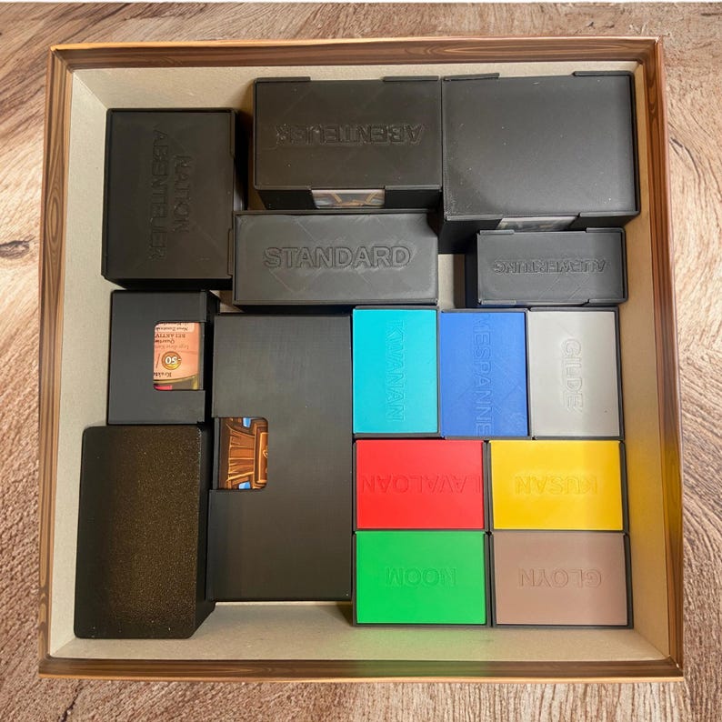 May include: A square board game box organizer containing multiple rectangular plastic containers in black, teal, blue, gray, red, yellow, green, and brown. The containers feature embossed text including Standard, Abenteuer, Gilde, and Gloyn.
