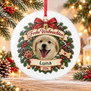 May include: A round white ceramic Christmas ornament featuring a golden retriever puppy portrait inside a wreath of pine branches, red berries, and pinecones. It includes a red bow, a gold bell, and banners with the text Frohe Weihnachten, Luna, and 2026.