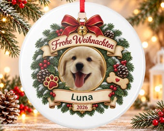May include: A round white ceramic Christmas ornament featuring a golden retriever puppy portrait inside a wreath of pine branches, red berries, and pinecones. It includes a red bow, a gold bell, and banners with the text Frohe Weihnachten, Luna, and 2026.