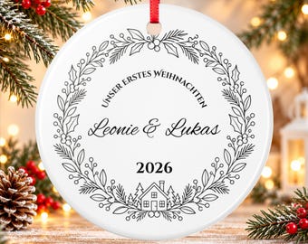 May include: A round white ceramic Christmas ornament with a red ribbon hanger. The design features a black line art wreath surrounding the text "UNSER ERSTES WEIHNACHTEN", the names "Leonie & Lukas", the year "2026", and a small house illustration.
