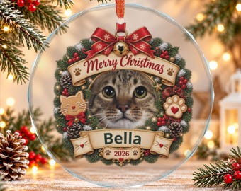 May include: A round, clear glass Christmas ornament featuring a brown tabby cat face centered within a festive wreath. The wreath includes red bows, pinecones, and berries. A banner reads Merry Christmas, Bella, and 2026.