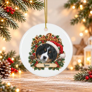 May include: A round white ceramic Christmas ornament featuring a Bernese Mountain Dog puppy wearing a Santa hat. The puppy is framed by a wreath of holly, pinecones, and red berries. The text Santa Paws is above, and the name Luna is below.