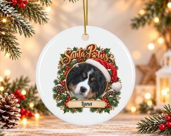 May include: A round white ceramic Christmas ornament featuring a Bernese Mountain Dog puppy wearing a Santa hat. The puppy is framed by a wreath of holly, pinecones, and red berries. The text Santa Paws is above, and the name Luna is below.