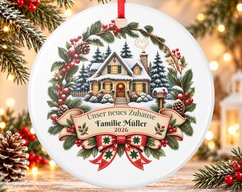 May include: A round white ceramic Christmas ornament featuring a snowy house scene surrounded by a wreath of pine branches and red berries. A banner at the bottom displays the text "Unser neues Zuhause", "Familie Müller", and "2026" above a red and green bow.