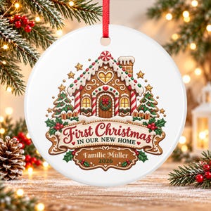 May include: A round white ceramic Christmas ornament featuring a gingerbread house illustration with a red ribbon hanger. The text reads First Christmas IN OUR NEW HOME, Familie Müller, and 2026. The design includes green pine trees and red berries.