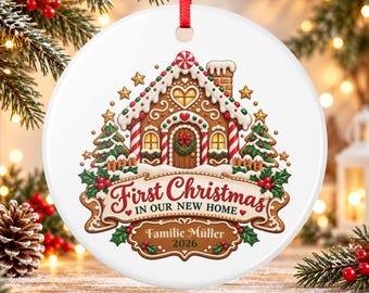 May include: A round white ceramic Christmas ornament featuring a gingerbread house illustration with a red ribbon hanger. The text reads First Christmas IN OUR NEW HOME, Familie Müller, and 2026. The design includes green pine trees and red berries.