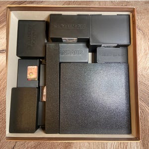 May include: A square board game box insert featuring multiple black plastic compartments for organizing game components. The boxes are labeled with embossed text including ABENTEUER, NATION ABENTEUER, STANDARD, and AUFENTHALT, with one large textured black tray.