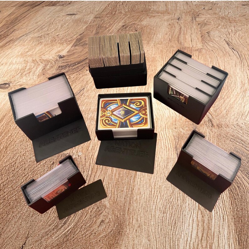 May include: Five black plastic card holders arranged on a surface. Four holders contain stacks of playing cards, while one central holder displays a single card with a blue, gold, and red ornate design. The holders feature embossed text.