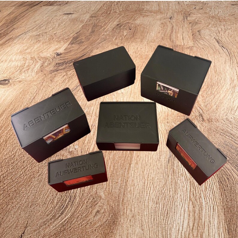 May include: Six black rectangular plastic storage boxes for game cards, arranged in a cluster. Some boxes feature embossed German text including ABENTEUER, NATION ABENTEUER, and AUFWERTUNG. Each box has a small cutout window revealing colorful card edges.