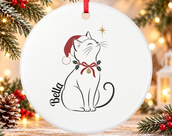 May include: A white ceramic round ornament featuring a black line drawing of a cat wearing a red Santa hat and a green holly collar with a gold bell. The name Bella is printed in black script next to the cat, with a gold star above.