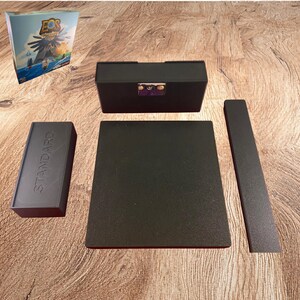 May include: A black plastic board game organizer set for Eos Island of Angels, including a rectangular card holder, a flat square tray, a long narrow tray, and a small rectangular box embossed with the word STANDARD on its top surface.