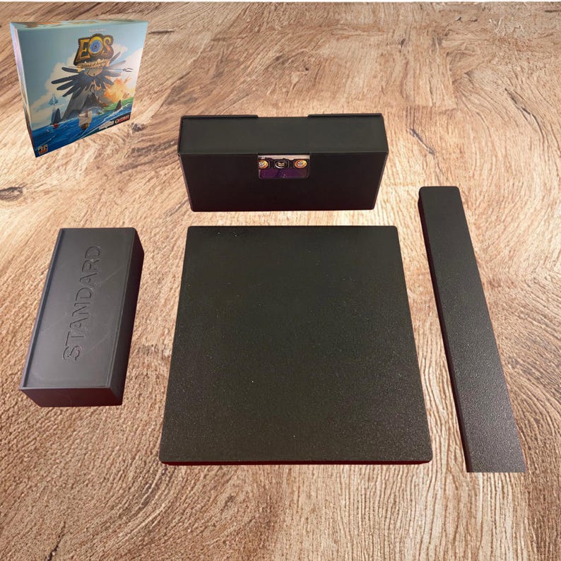 May include: A black plastic board game organizer set for Eos Island of Angels, including a rectangular card holder, a flat square tray, a long narrow tray, and a small rectangular box embossed with the word STANDARD on its top surface.