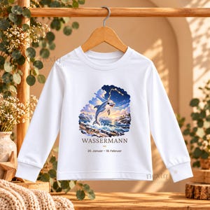 May include: A white long-sleeve shirt featuring a graphic of a dolphin leaping from ocean waves under a starry sky with a lighthouse. Below the graphic, the text reads WASSERMANN and 20. Januar - 18. Februar in black lettering.
