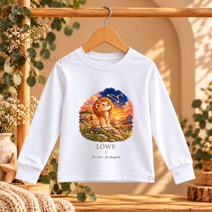 May include: A white long-sleeve shirt featuring a graphic of a cartoon lion sitting on a rock at sunset. Below the illustration, the text reads L&Ouml;WE, the Leo zodiac symbol, and the date range 23. Juli &ndash; 22. August in black lettering.
