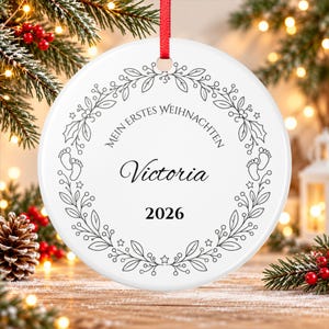 May include: A round white ceramic ornament with a red ribbon hanger. The design features a black line art wreath of leaves, berries, and baby footprints. Text reads: MEIN ERSTES WEIHNACHTEN, the name Victoria, and the year 2026 in black font.