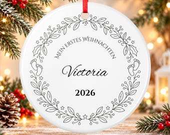 May include: A round white ceramic ornament with a red ribbon hanger. The design features a black line art wreath of leaves, berries, and baby footprints. Text reads: MEIN ERSTES WEIHNACHTEN, the name Victoria, and the year 2026 in black font.