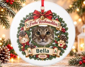 May include: A round white ceramic Christmas ornament featuring a brown tabby cat portrait centered in a festive wreath. The design includes a red bow, pinecones, holly berries, a small cat illustration, a paw print, and the text Frohe Weihnachten, Bella, and 2026.
