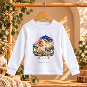 K&ouml;nnte beinhalten: Ein wei&szlig;es Langarmshirt mit einer Grafik eines Widders auf einem felsigen H&uuml;gel mit Blumen, Bergen und einem Sternenhimmel. Unter der Illustration steht der Text WIDDER, das Widder-Symbol und 21. M&auml;rz &ndash; 19. April.