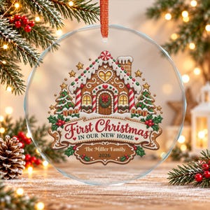 May include: A round, clear glass ornament with a red ribbon hanger featuring a gingerbread house illustration, green pine trees, and red holly berries. The text reads First Christmas IN OUR NEW HOME, The Miller Family, and 2026.