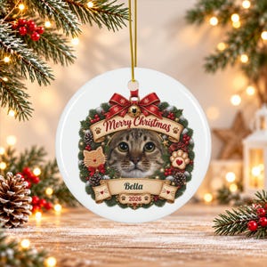 May include: A round white ceramic Christmas ornament featuring a central portrait of a tabby cat framed by a festive wreath of pine, red berries, and pinecones. The design includes a red bow, the text Merry Christmas, Bella, and 2026.