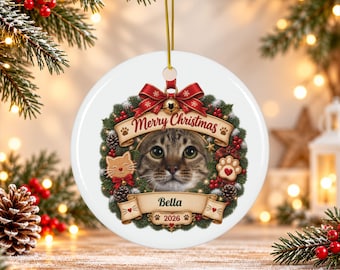 May include: A round white ceramic Christmas ornament featuring a central portrait of a tabby cat framed by a festive wreath of pine, red berries, and pinecones. The design includes a red bow, the text Merry Christmas, Bella, and 2026.