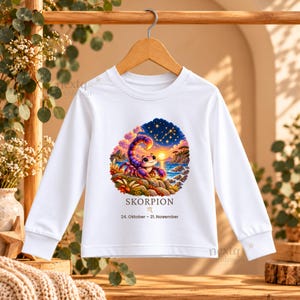 Puede incluir: Una camiseta blanca de manga larga con un gr&aacute;fico colorido de un escorpi&oacute;n de dibujos animados en una costa rocosa al atardecer, con el texto SKORPION, el s&iacute;mbolo del zodiaco Escorpio y las fechas 24. Oktober - 21. November.
