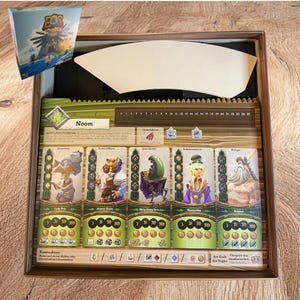 May include: A board game box containing a game board with the title Noom, featuring five character cards with illustrations of fantasy figures, a numbered track from 0 to 25, and various icons. A separate game box lid is shown in the top left corner.