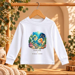 Puede incluir: Una camiseta blanca de manga larga con un gr&aacute;fico submarino colorido de dos peces, coral y una estrella de mar. Debajo de la ilustraci&oacute;n, el texto dice FISCHE, el s&iacute;mbolo del zodiaco Piscis y 19. Februar &ndash; 20. M&auml;rz.
