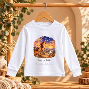 May include: A white long-sleeve shirt featuring a graphic of a cartoon centaur standing on a rocky cliff at sunset. Above the centaur is a constellation. Below the graphic is the text SCH&Uuml;TZE and 22. November - 21. Dezember.