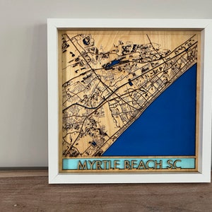 May include: A white framed wooden map of Myrtle Beach, South Carolina. The map is laser cut and features a blue ocean and a light blue border with the text "Myrtle Beach SC".