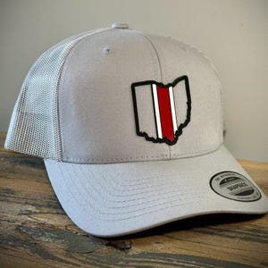 May include: Gray trucker hat with a mesh back and a curved brim. The hat features a patch of the Ohio state outline in black with a red and white stripe. The hat has a Snapback tag.